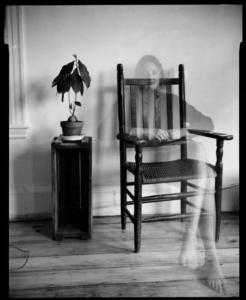 A ghostly figure of a woman sits on a wooden slat chair in a room with a plan on a stand.
