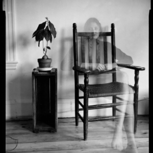 a ghostly figure of a woman sitting on a slat-back chair. Photo by Susan Meiselas