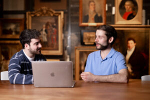 Two students at a table with laptop in front of paintings